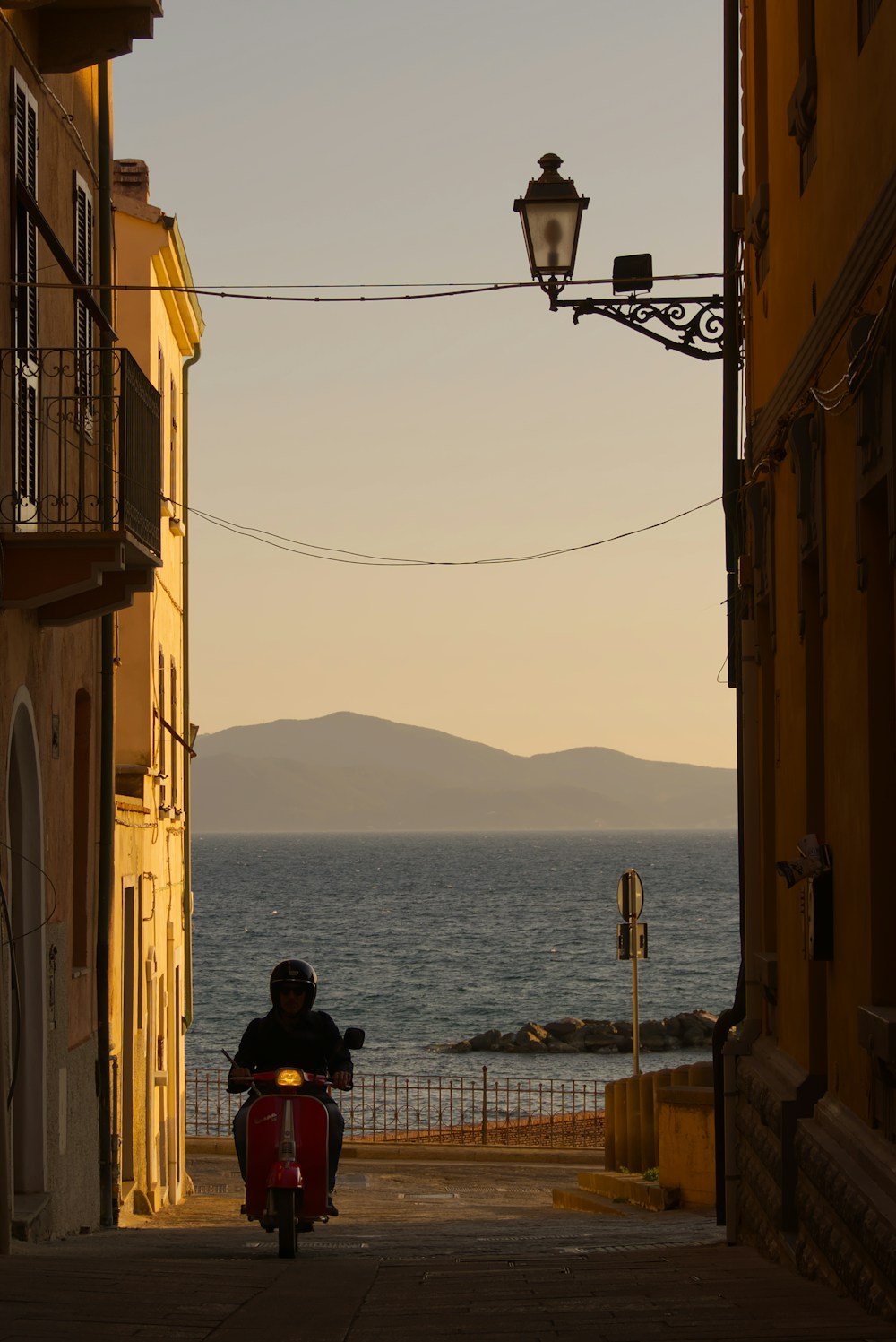 Person on red scooter riding toward the sea at golden hour